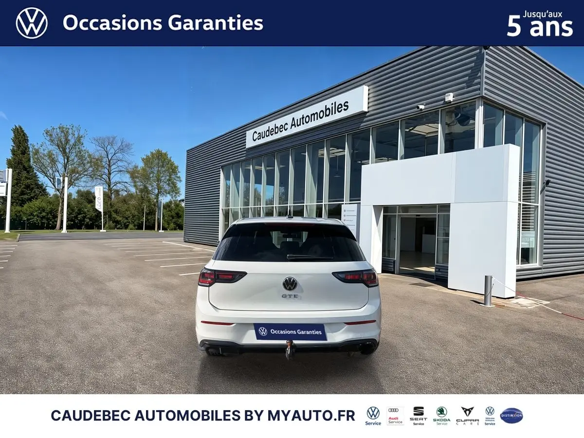 Vue arrière d'une Volkswagen Golf 1.5 eHybrid blanche avec attelage électrique, stationnée devant un bâtiment moderne.