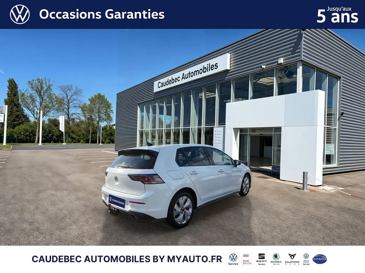 Vue 3/4 arrière droite d'une Volkswagen Golf 1.5 eHybrid blanche stationnée devant un bâtiment moderne de concession automobile.