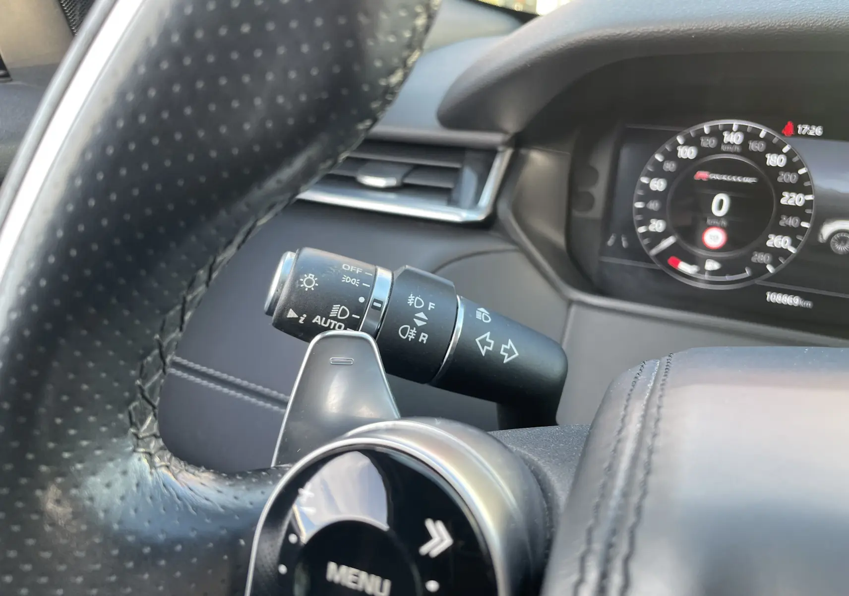 Gros plan sur le volant en cuir perforé et la commande des phares du Range Rover Velar noir Santorini, tableau de bord digital visible.