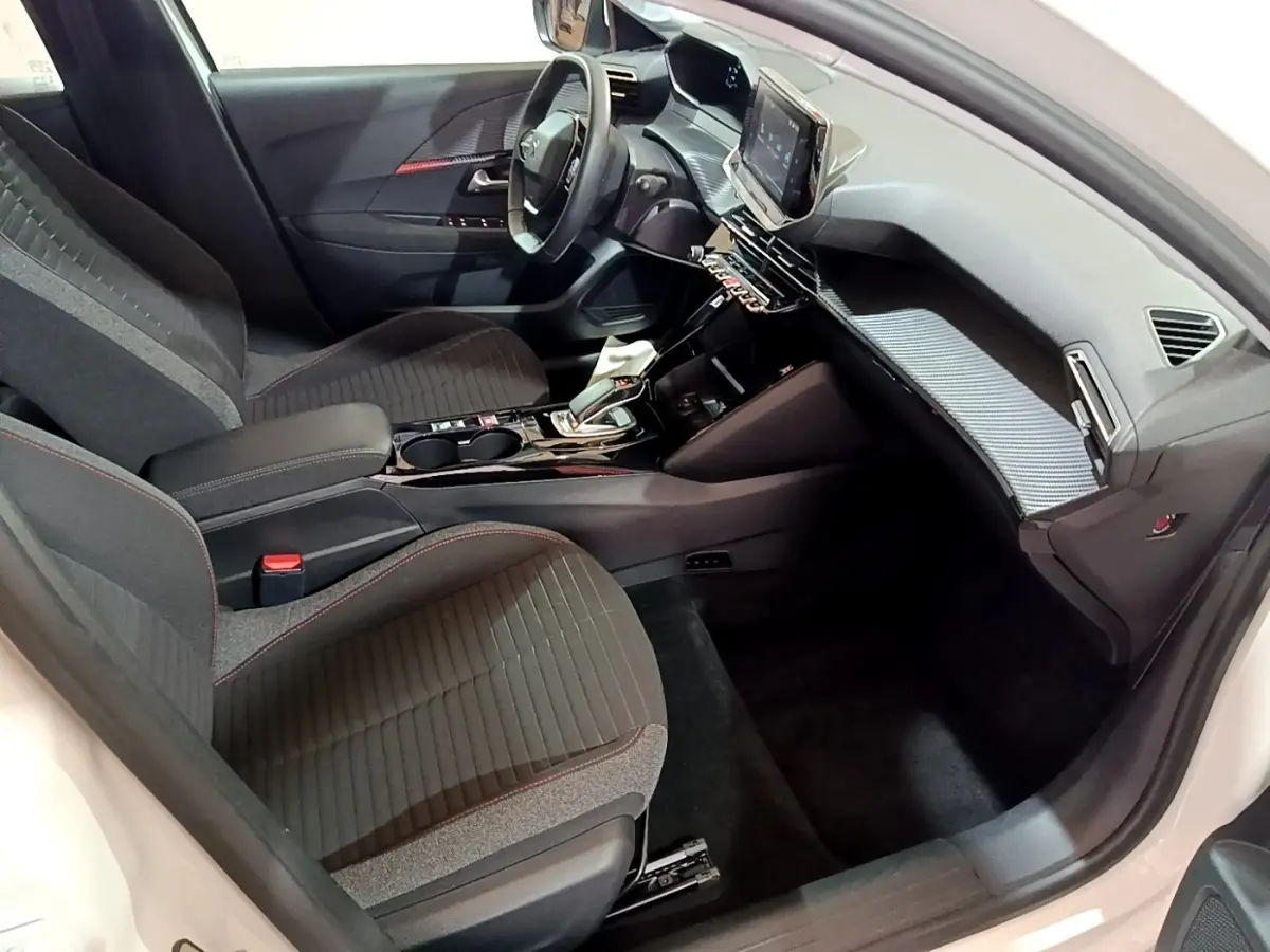 Intérieur avant droit de la Peugeot 208 électrique blanche, avec tableau de bord moderne et sièges gris à surpiqûres rouges.