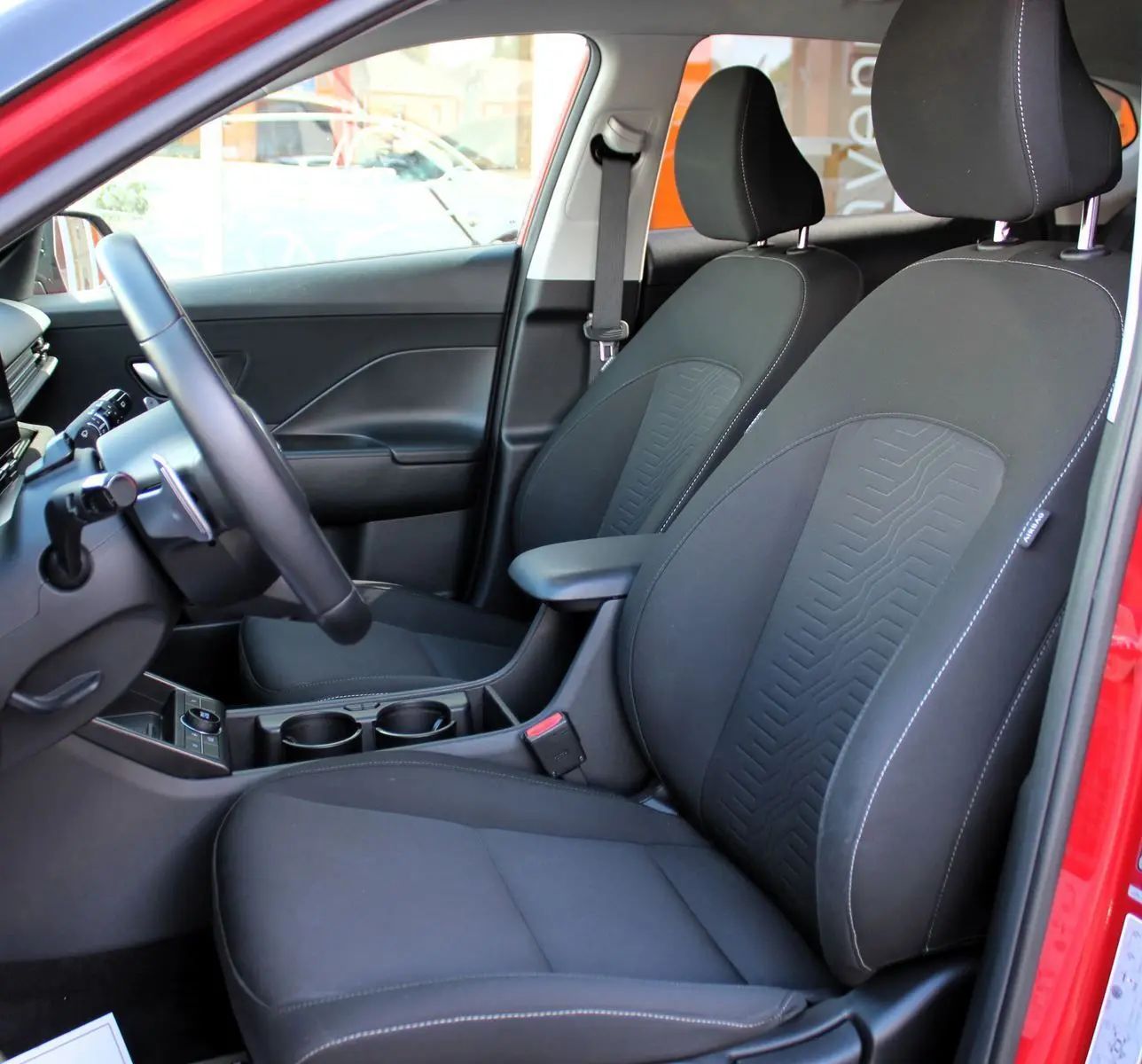 Vue intérieure côté conducteur du Hyundai Kona 2024 rouge, sièges avant noirs en tissu avec surpiqûres blanches.