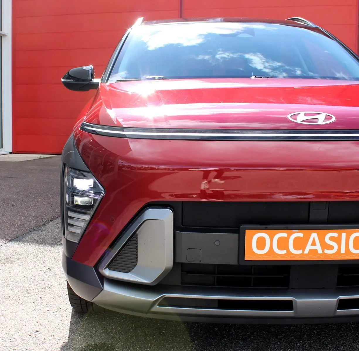 Vue avant rapprochée d’un Hyundai Kona rouge Ultimate Metal avec calandre noire et détails gris, devant un mur rouge.