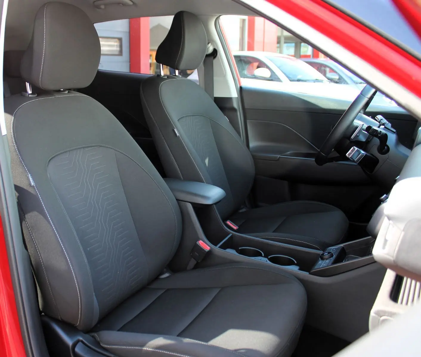 Vue intérieure côté gauche du Hyundai Kona 2024 hybride rouge, montrant les sièges avant en tissu noir avec accoudoir central.