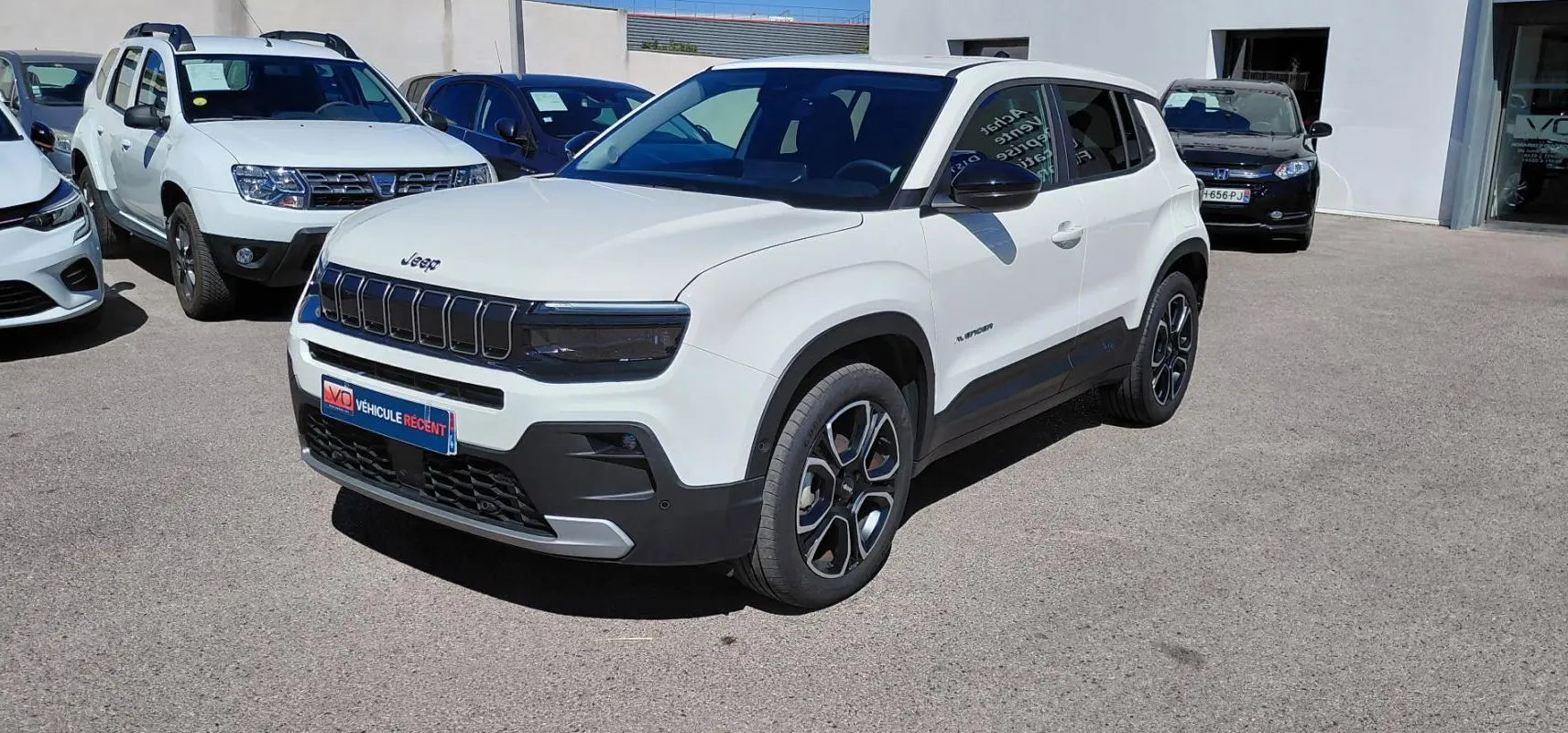 Jeep Avenger blanc en 3/4 avant droit, avec calandre noire et jantes distinctives, stationné en extérieur.