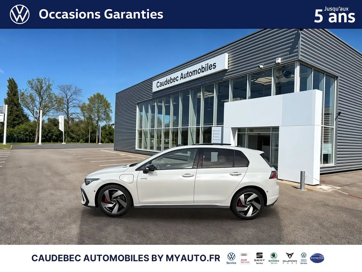Vue de profil côté gauche d'une Volkswagen Golf 1.5 eHybrid blanche stationnée devant un bâtiment moderne en verre.