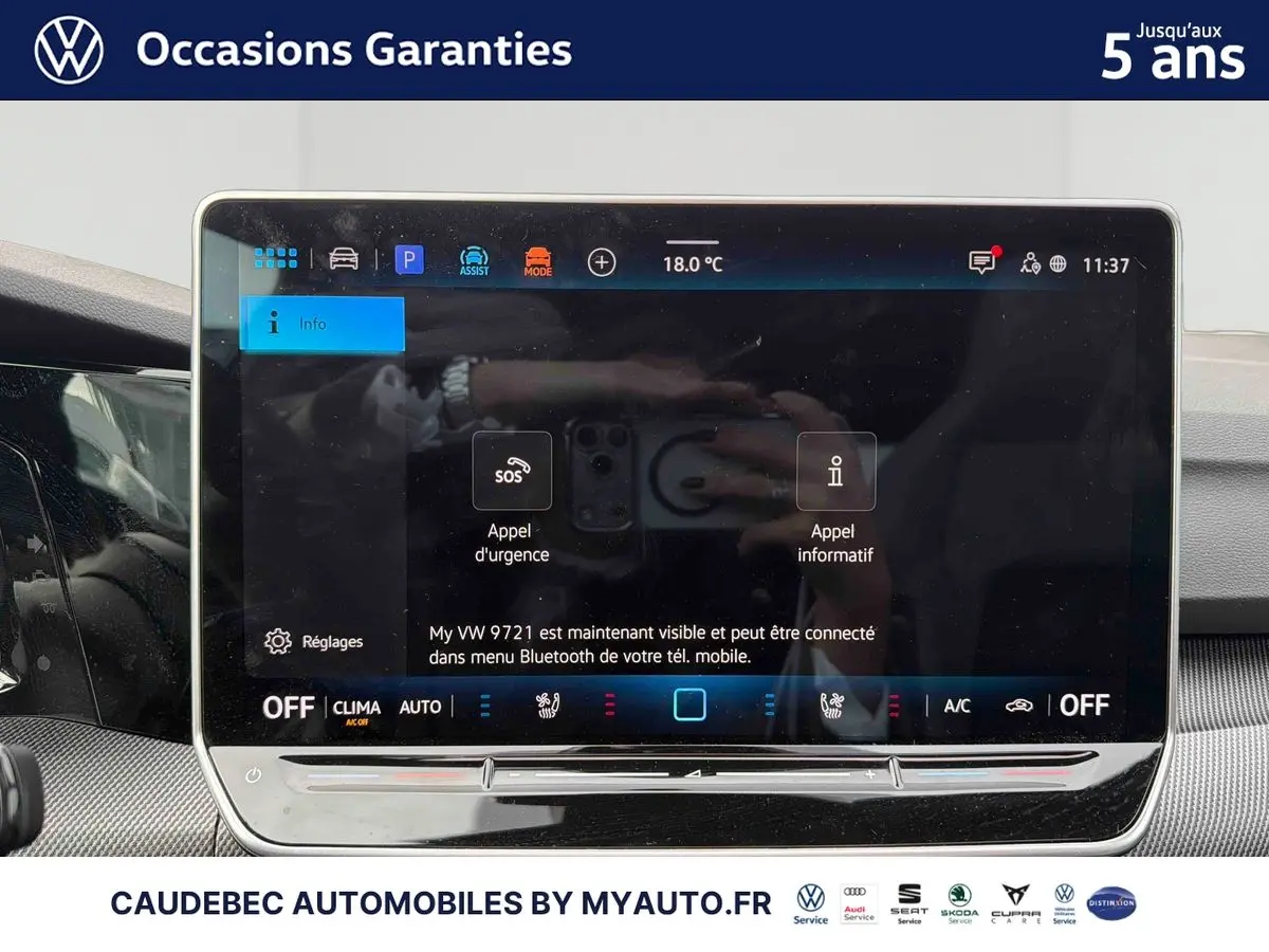 Vue intérieure centrée sur l'écran tactile du tableau de bord de la Volkswagen Golf blanche, affichant les commandes d'urgence et climatisation.
