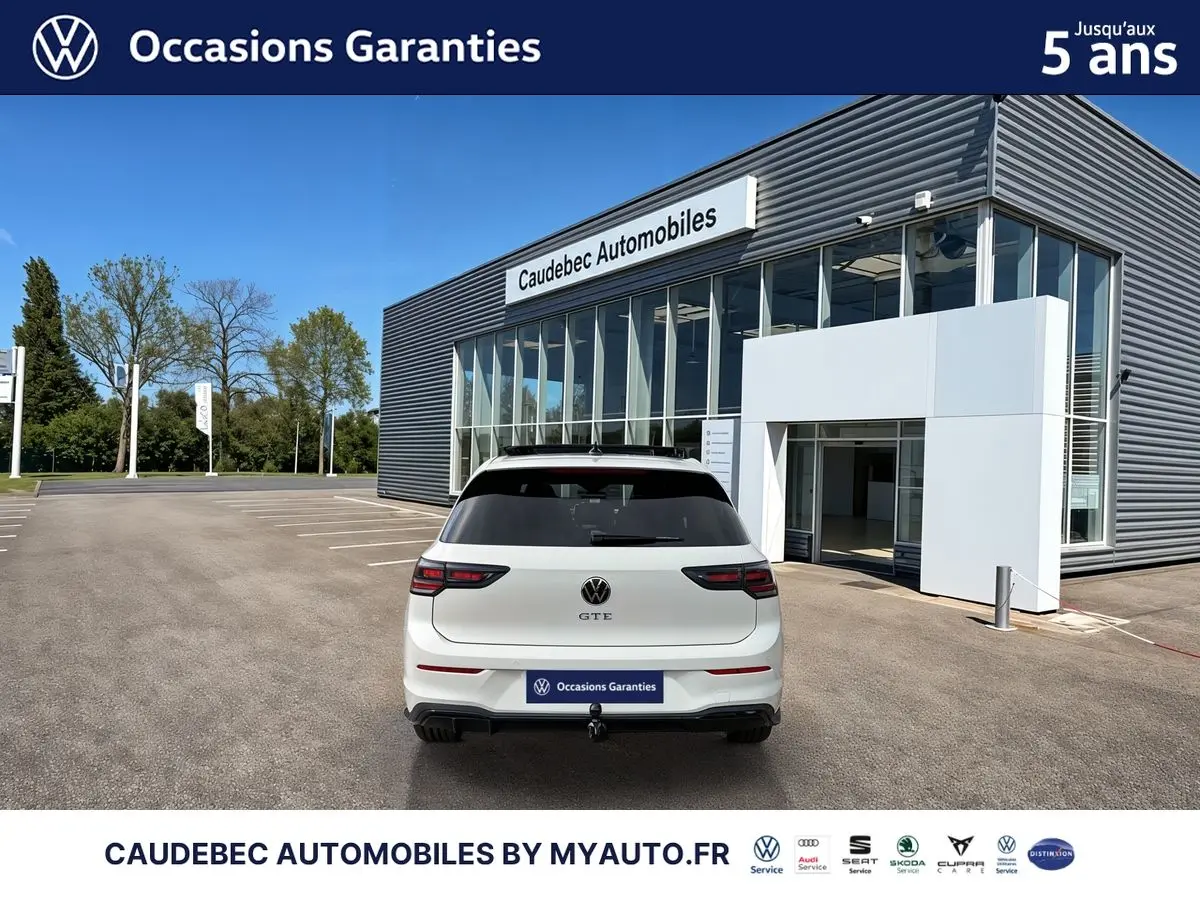 Vue arrière d'une Volkswagen Golf 1.5 eHybrid blanche, avec attelage escamotable, devant un bâtiment de concession.