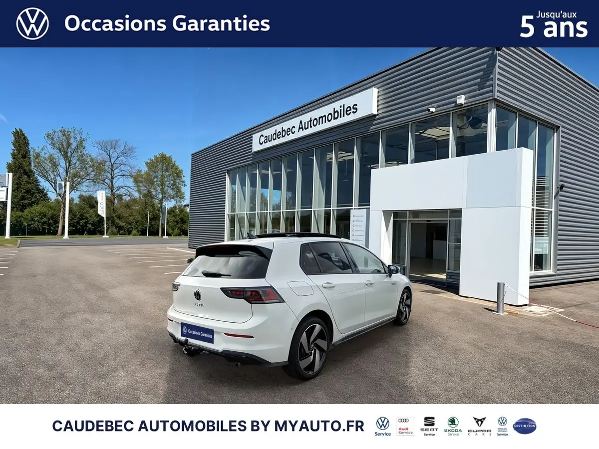 Vue 3/4 arrière droite d'une Volkswagen Golf 1.5 eHybrid blanche nacrée, stationnée devant un bâtiment moderne.