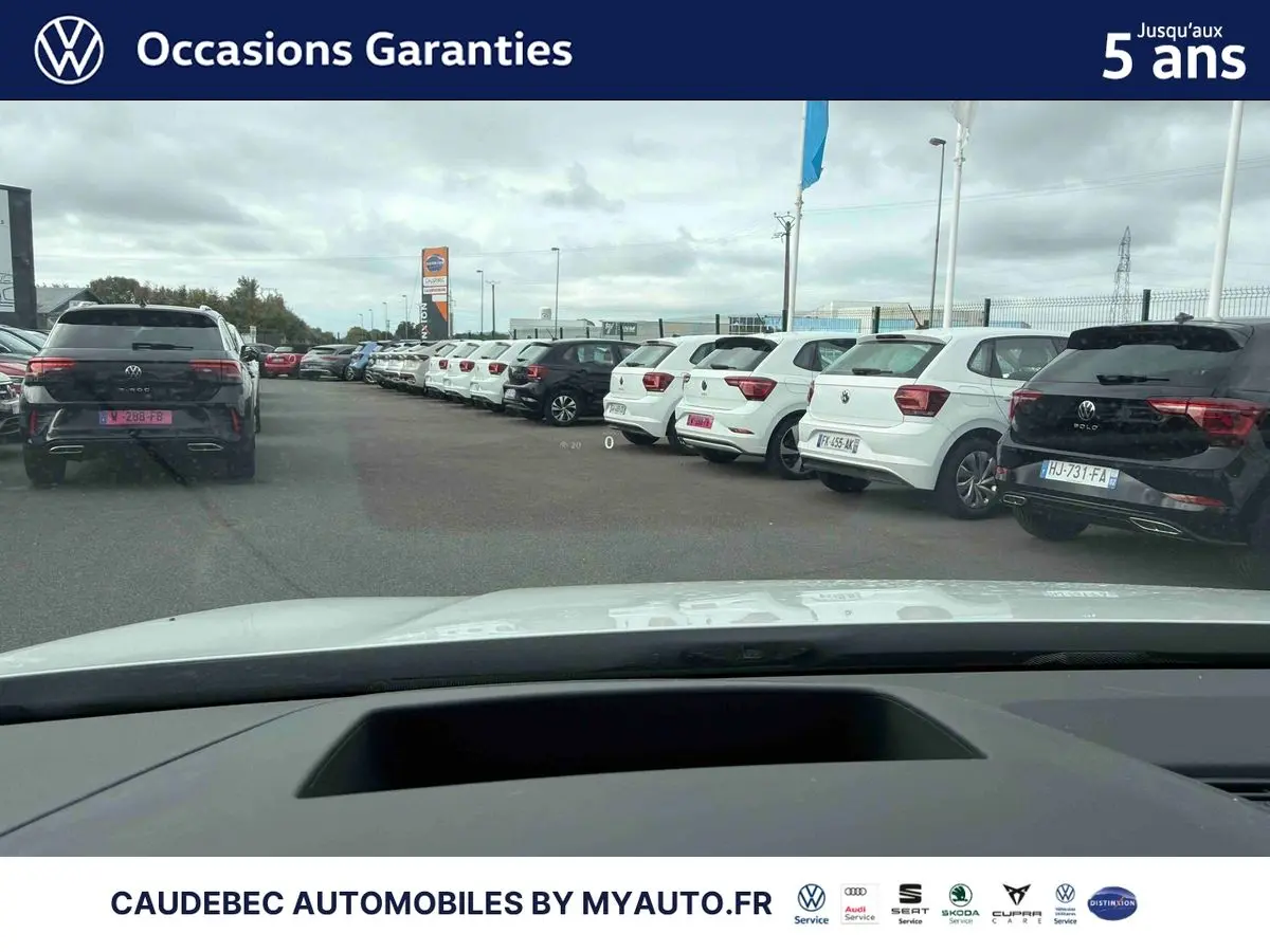 Vue depuis l’intérieur d’une Volkswagen Golf blanche, capot visible, sur un parking avec plusieurs voitures blanches et noires alignées.