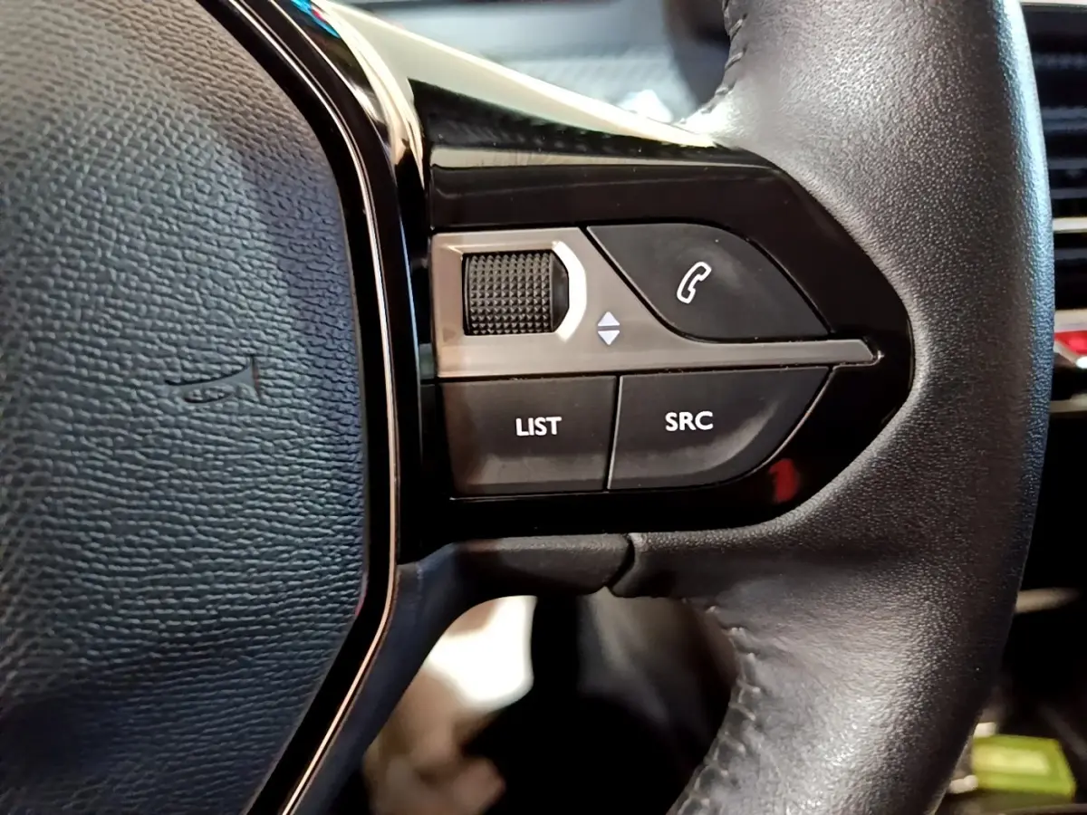 Gros plan sur les commandes gauche du volant cuir noir du Peugeot 208 électrique blanc, boutons de gestion audio et appel.