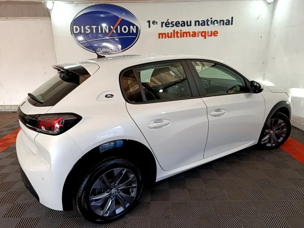 Vue 3/4 arrière droite d'une Peugeot 208 électrique Active Business blanche en intérieur, avec jantes foncées et logo "e" sur l'aile arrière.