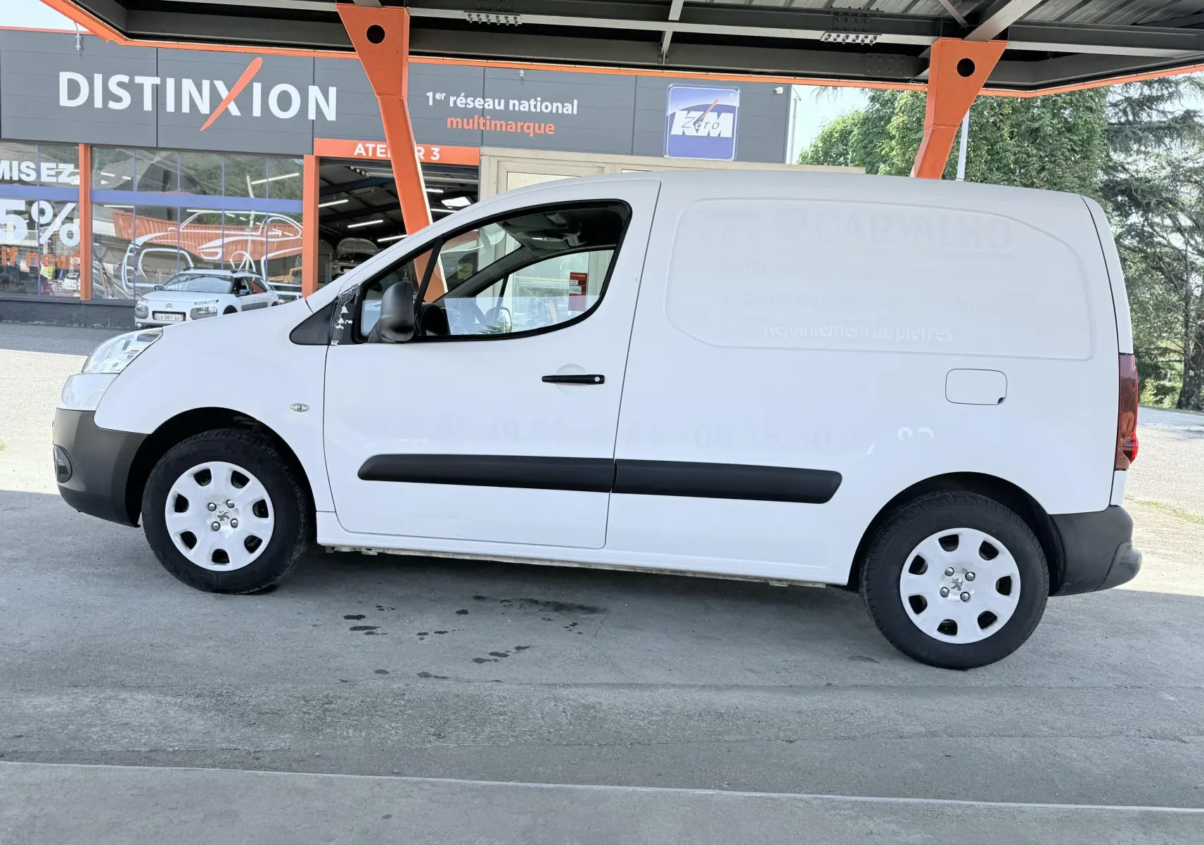 Vue de profil côté gauche d’un utilitaire Peugeot Partner blanc, avec bandes de protection noires et jantes acier sous un abri.