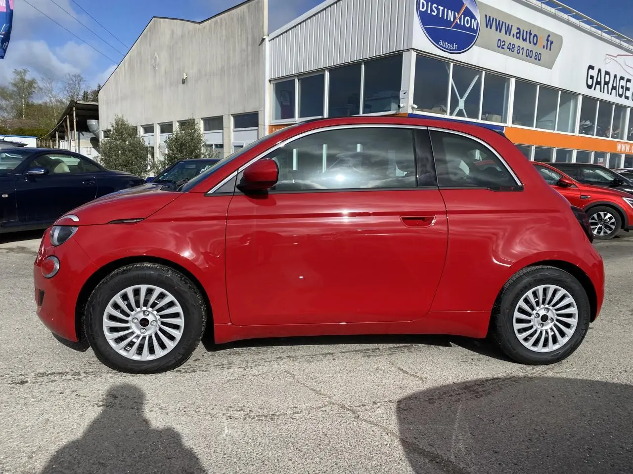 Profil latéral gauche d'une FIAT 500 Cabriolet Electrique rouge stationnée devant un garage avec jantes alliage 16 pouces.