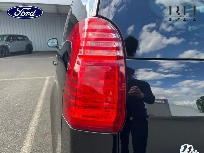 Gros plan sur le feu arrière gauche d’un Peugeot 5008 noir Perla Nera, reflétant le ciel et un photographe.