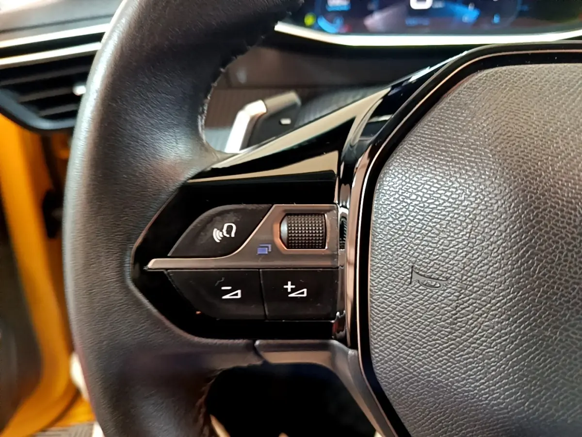 Gros plan sur le volant cuir de la Peugeot 208 jaune, avec commandes multifonctions et tableau de bord digital en arrière-plan.
