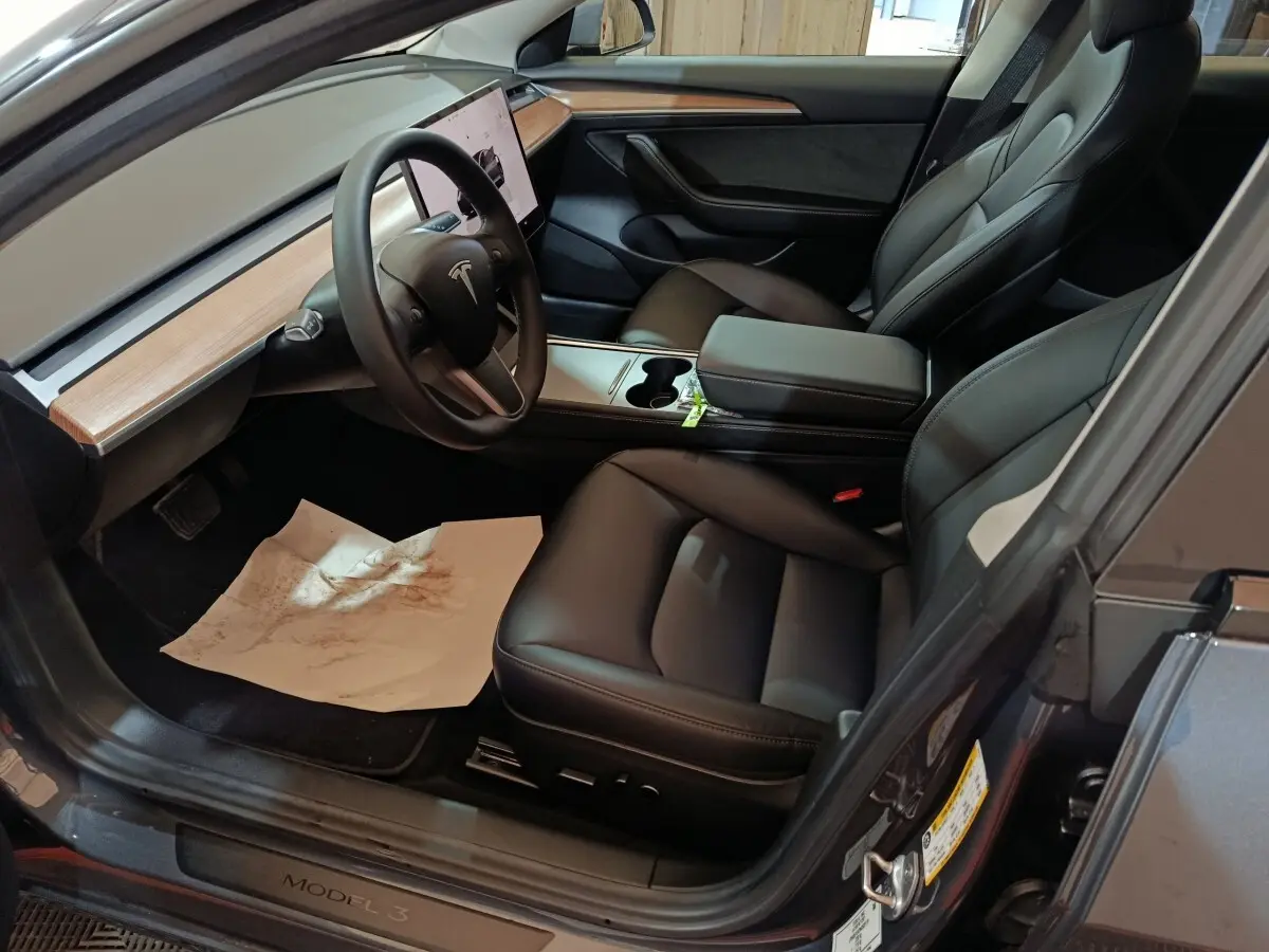 Intérieur Tesla Model 3 gris foncé, vue côté conducteur, sièges cuir noir et tableau de bord bois clair.