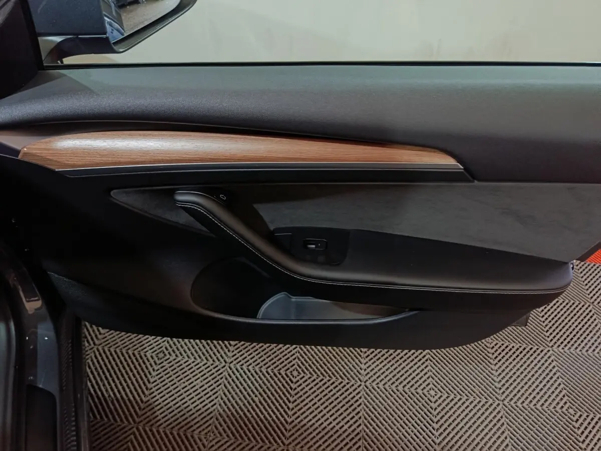 Vue intérieure du panneau de porte côté gauche de la Tesla Model 3 gris foncé, avec insert bois et poignée noire.