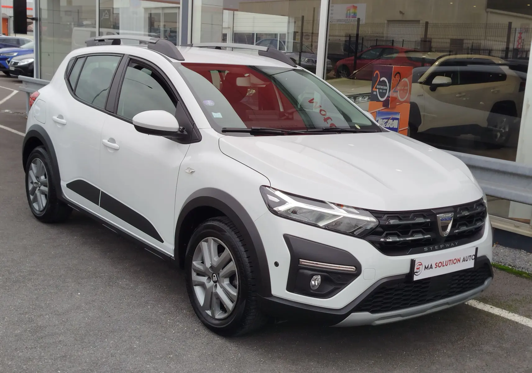 Vue 3/4 avant d'une Dacia Sandero Stepway blanche glacier avec détails noirs et barres de toit grises.