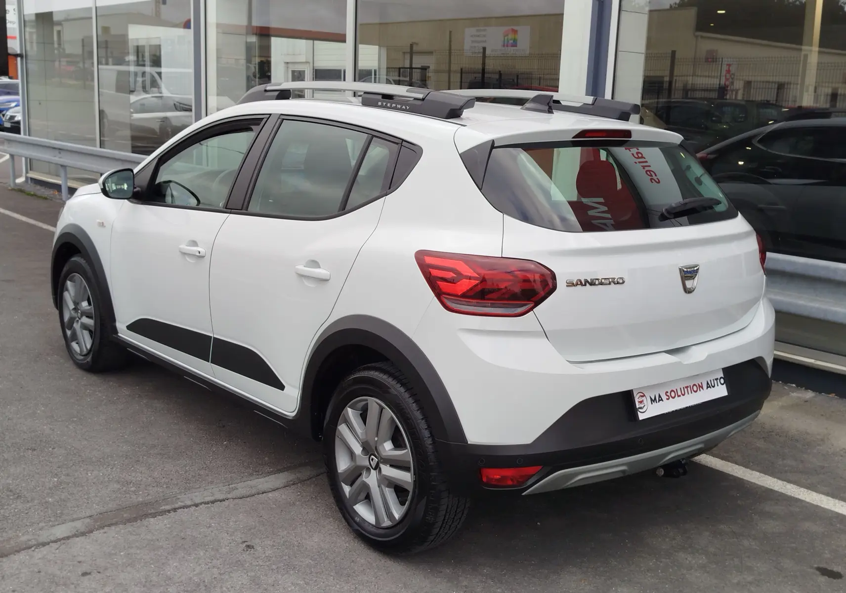 Vue 3/4 arrière droite d'une Dacia Sandero Stepway blanche Glacier avec barres de toit et protections noires.