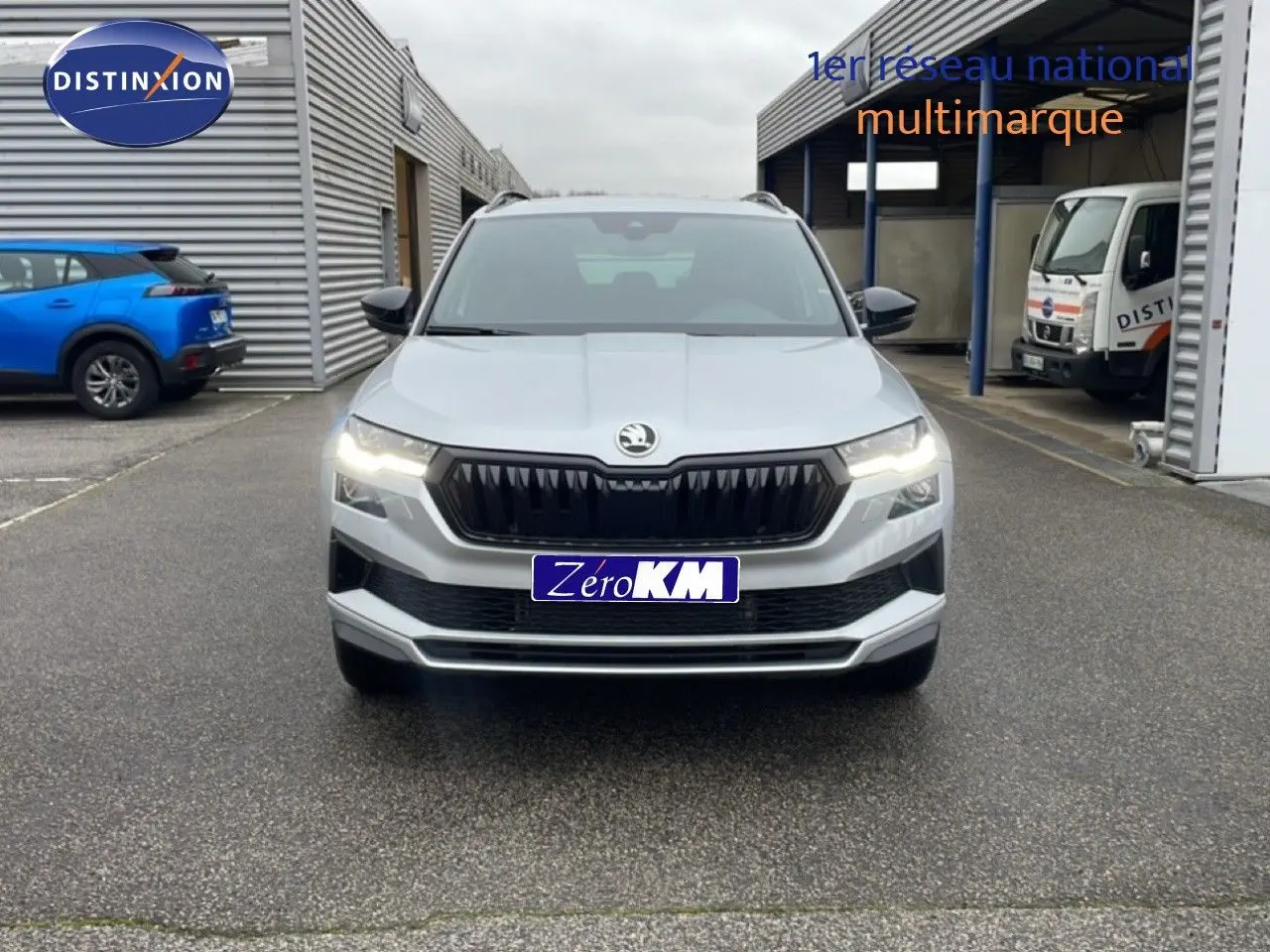 Vue frontale d'un Skoda Karoq gris argent métallisé 2025 avec calandre noire et phares allumés sur parking.