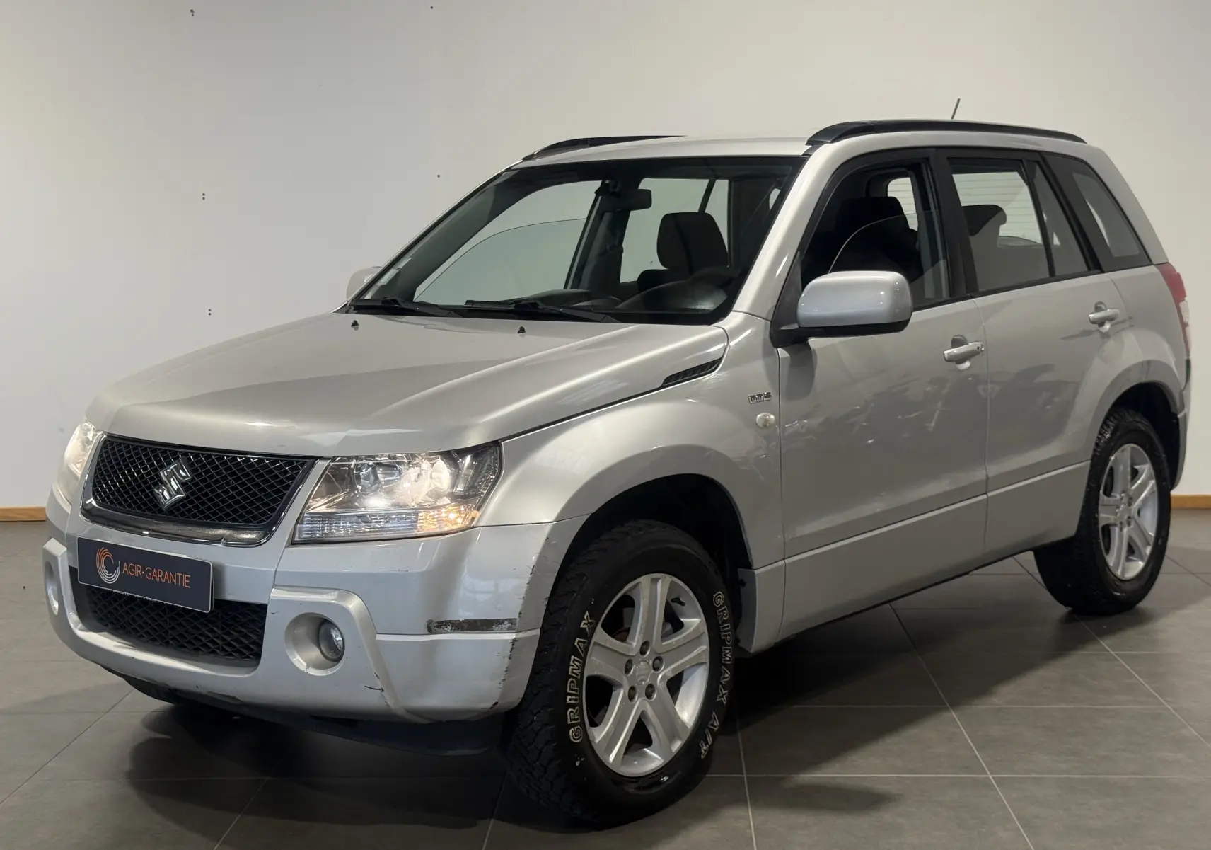 Suzuki Grand Vitara gris en 3/4 avant droit, SUV 2007 avec jantes alliage et barres de toit visibles.