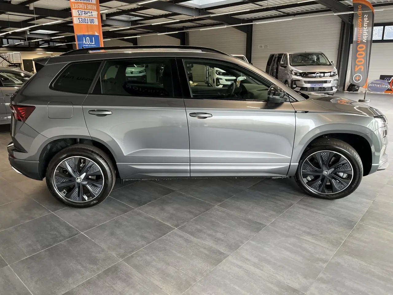 Profil côté gauche d'un SUV SKODA KAROQ Sportline gris perle avec jantes alliage noires et toit noir en intérieur showroom.