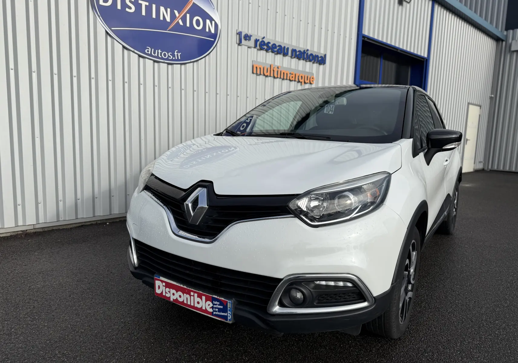 Renault Captur blanc en 3/4 avant droit, avec calandre noire et feux LED, stationné devant un bâtiment industriel.