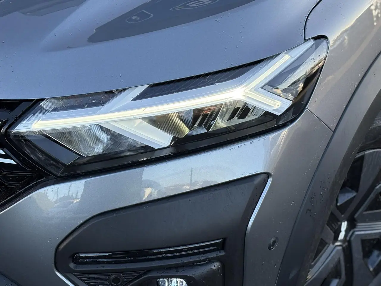 Gros plan sur le phare avant droit allumé du Dacia Jogger Gris Schiste avec enjoliveur noir et jante 16 pouces.