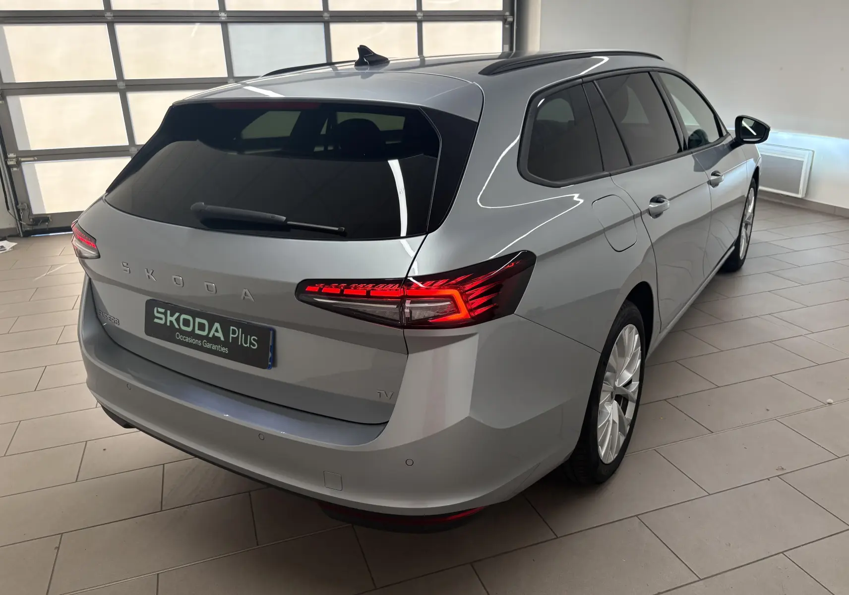 Vue 3/4 arrière droite d'une Skoda Superb Combi gris argent avec feux arrière LED allumés en intérieur showroom.