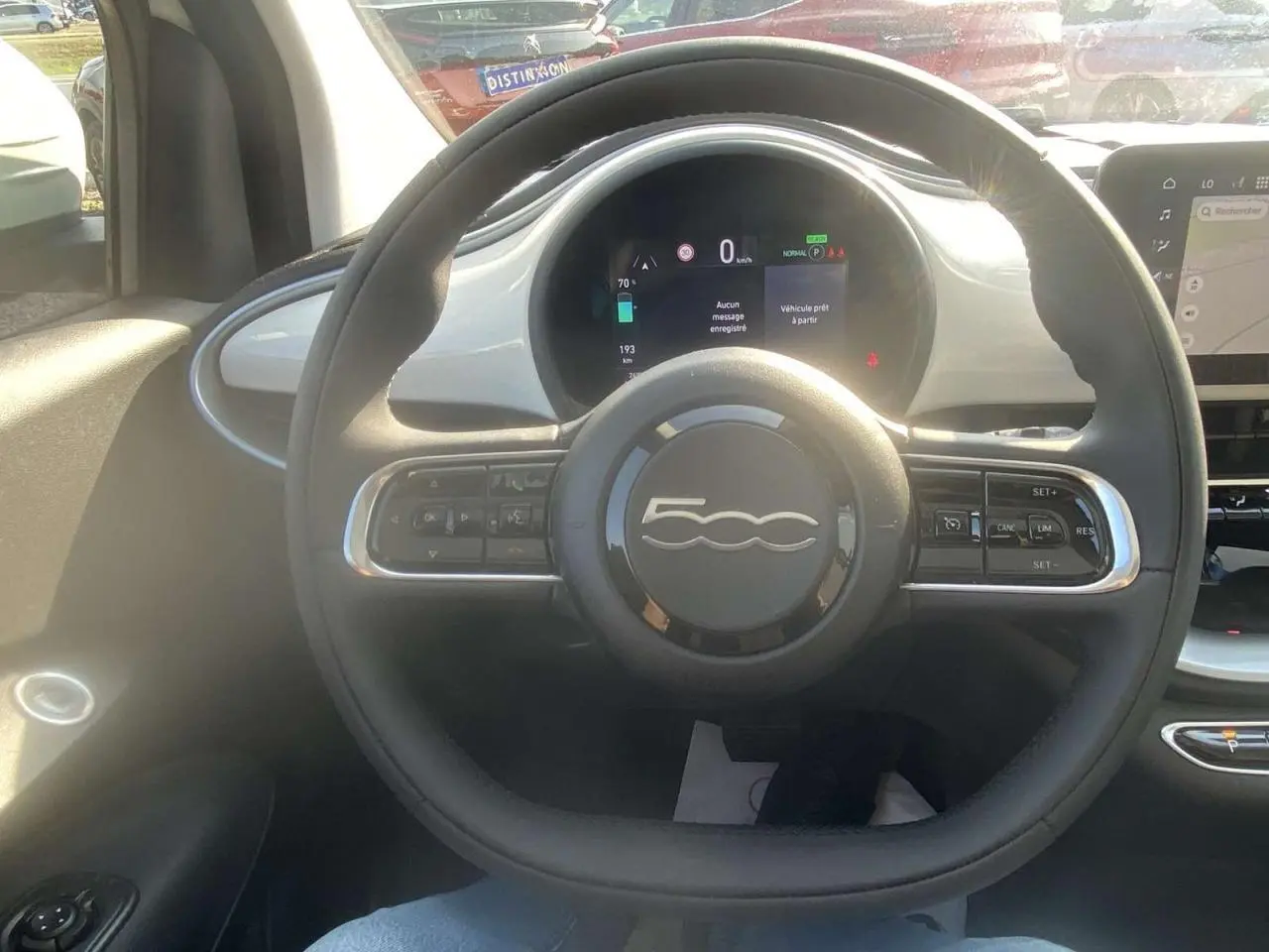 Vue intérieure centrée sur le volant noir de la Fiat 500 électrique blanche avec tableau de bord numérique lumineux.
