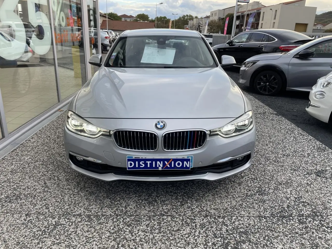 Vue frontale d'une BMW Série 3 F30 gris clair avec calandre chromée et phares LED allumés sur parking.
