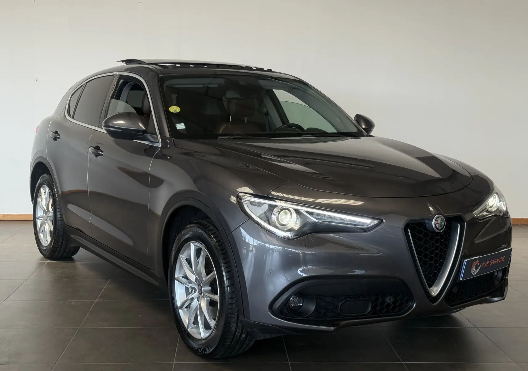 Alfa Romeo Stelvio gris en 3/4 avant droit, avec calandre caractéristique en V et phares allumés.