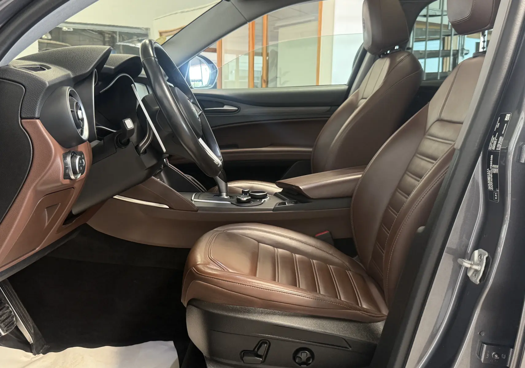 Intérieur de l'Alfa Romeo Stelvio 2017 vu côté conducteur, sièges en cuir marron et tableau de bord noir-gris.