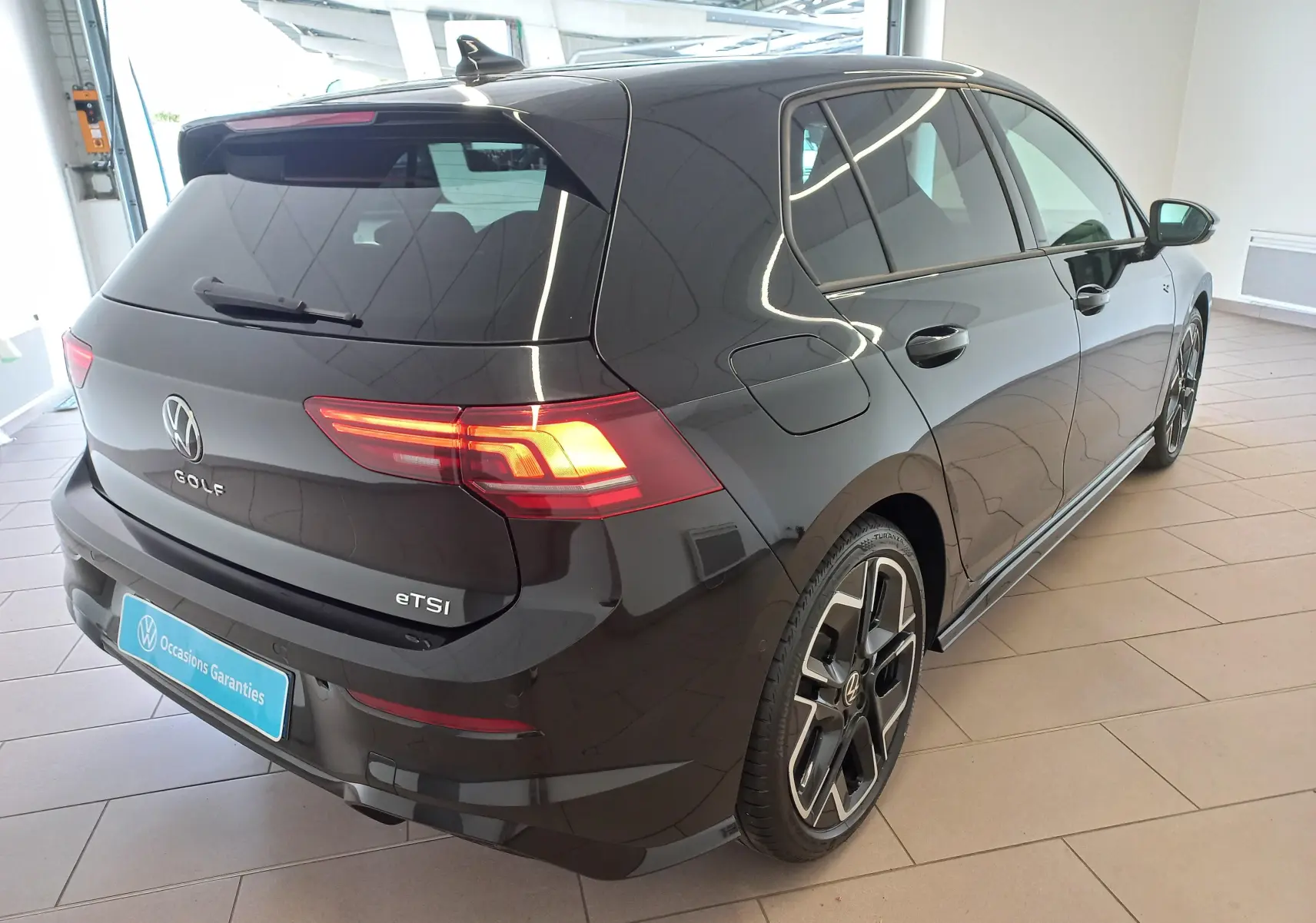 Vue 3/4 arrière droite d'une Volkswagen Golf 1.5 eTSI 150 DSG7 R-Line Edition noire Ébène avec feux allumés en intérieur showroom.