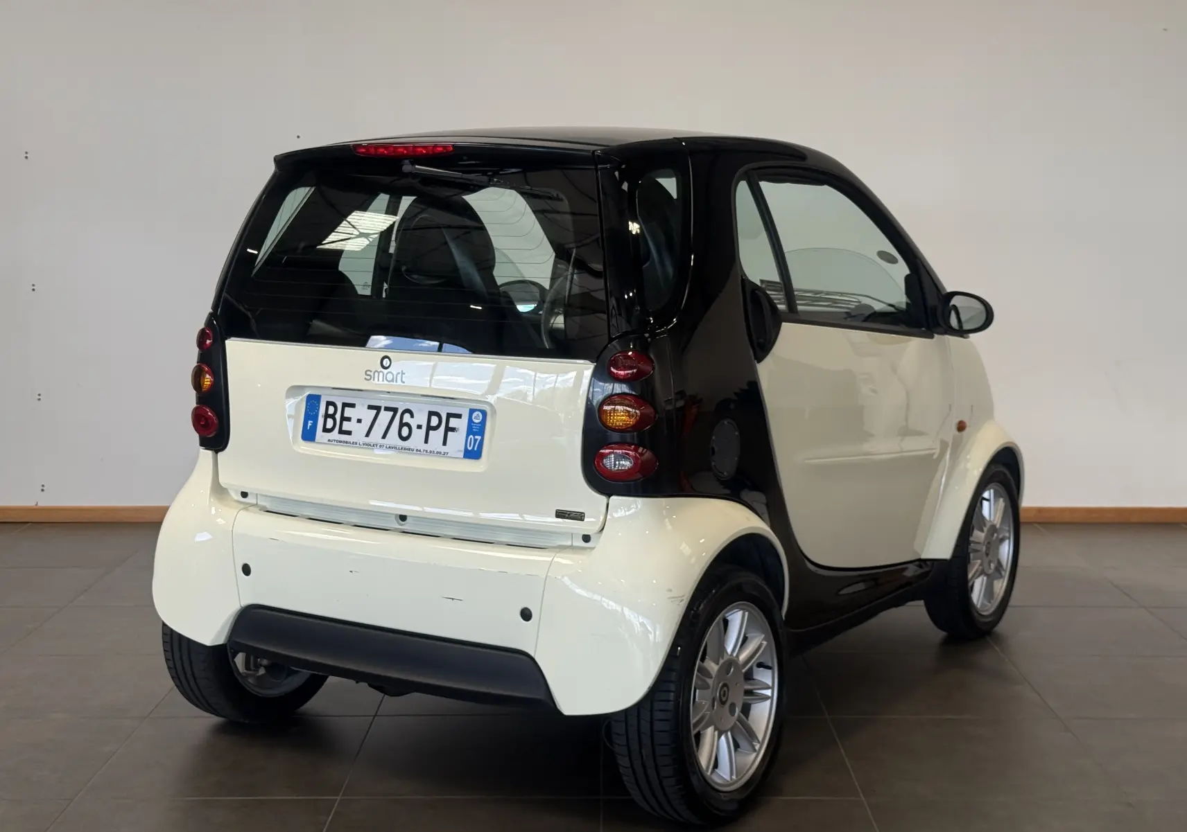 Vue 3/4 arrière droite d'une Smart Fortwo Coupé beige et noire de 2004 avec toit panoramique en intérieur showroom.