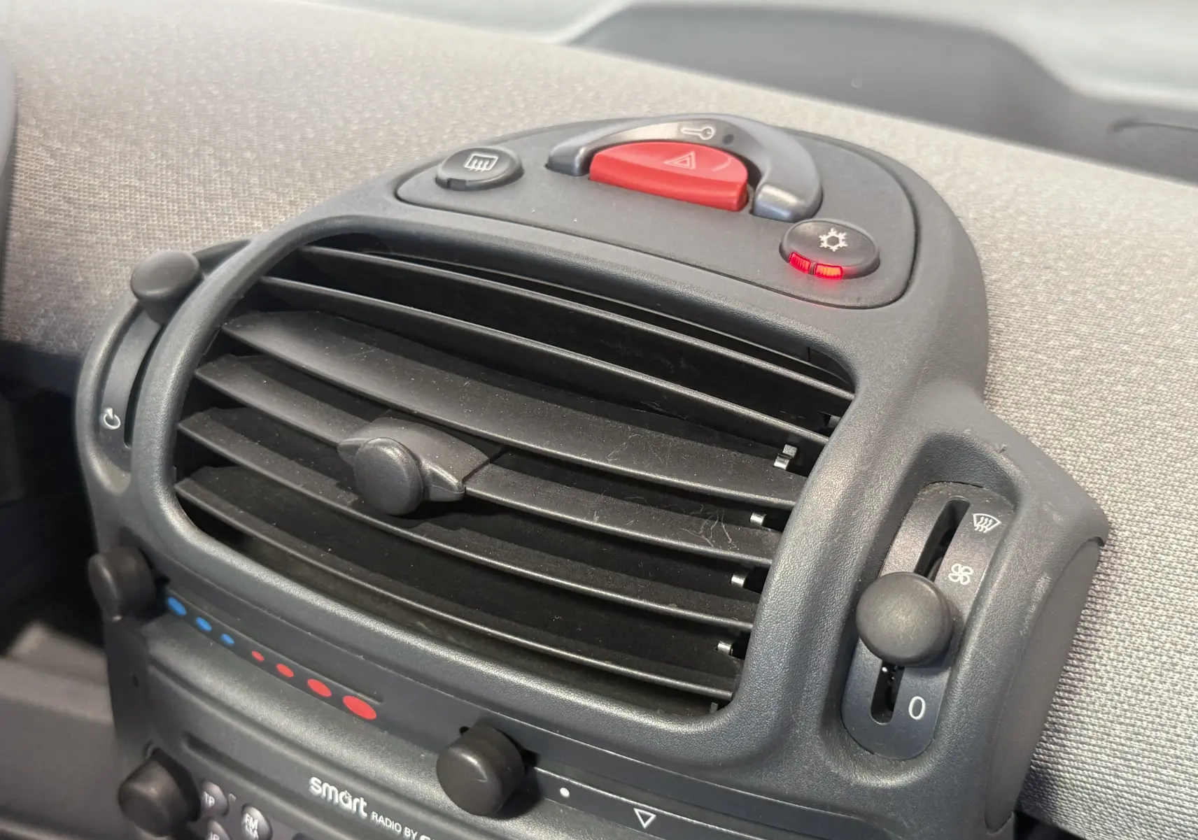Gros plan sur la console centrale noire avec commandes de ventilation et bouton d'alerte de la Smart Fortwo beige 2004.