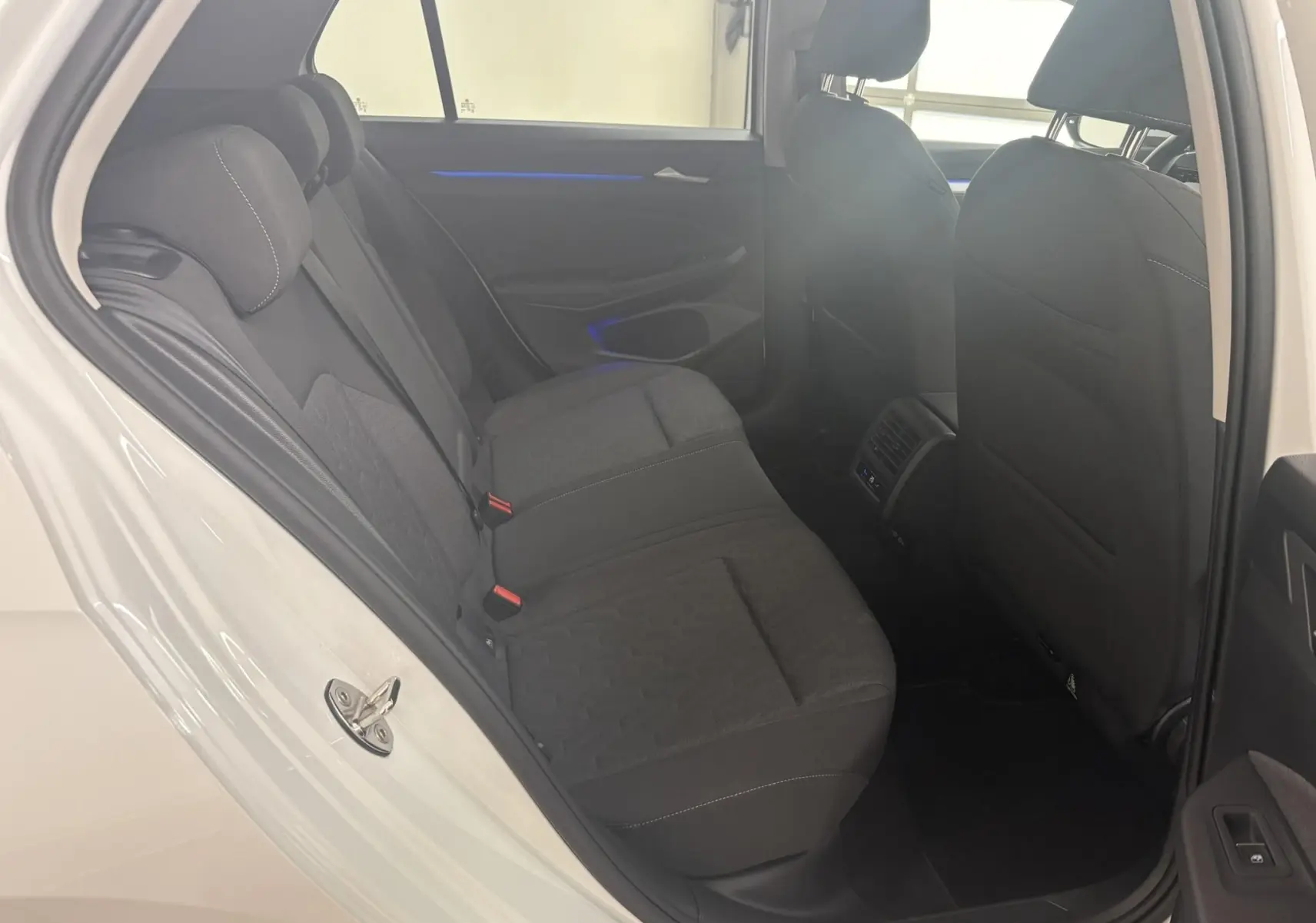 Vue intérieure arrière côté gauche d'une Volkswagen Golf 1.5 eTSI 2025 en blanc Oryx nacré, sièges tissu noirs et éclairage d'ambiance bleu.