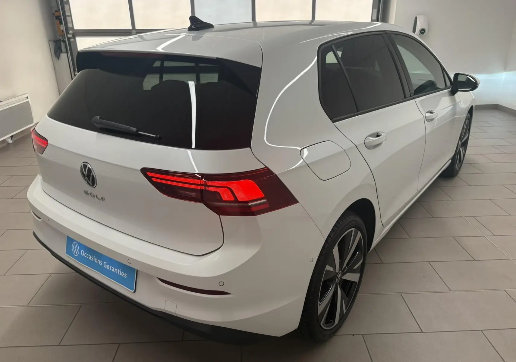 Vue 3/4 arrière droite d'une Volkswagen Golf 1.5 eTSI blanche nacrée avec jantes alliage 18'' Catania en intérieur showroom.