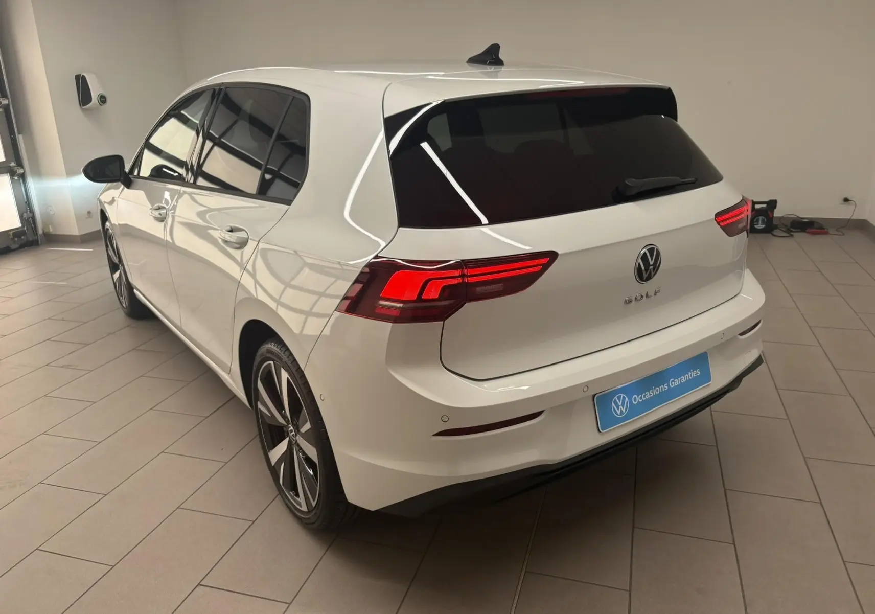 Vue 3/4 arrière droite d'une Volkswagen Golf 1.5 eTSI blanche nacrée avec jantes alliage 18'' Catania en intérieur showroom.