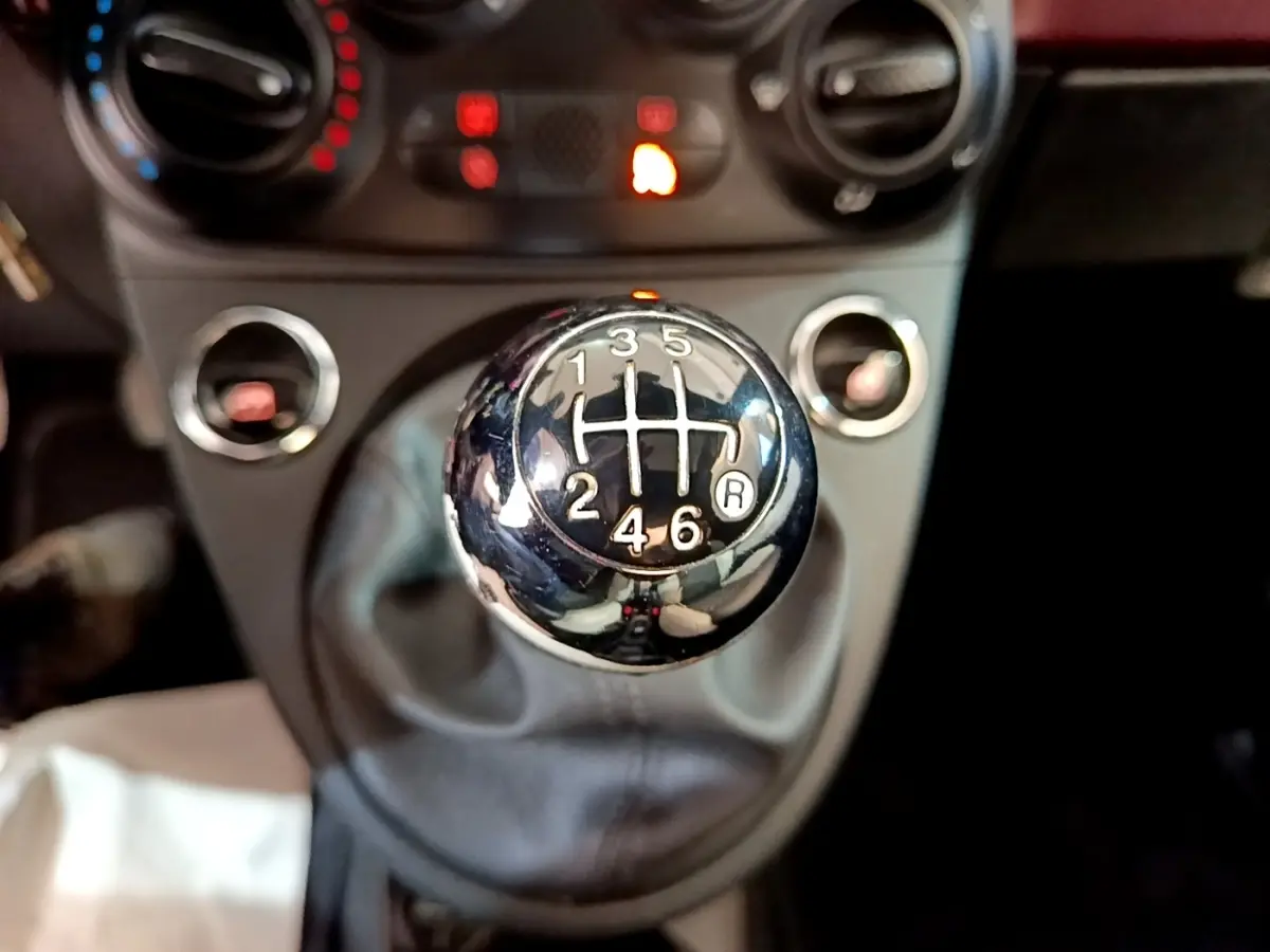 Gros plan sur le levier de vitesses manuel chromé de la FIAT 500 rouge, avec tableau de bord flou en arrière-plan.