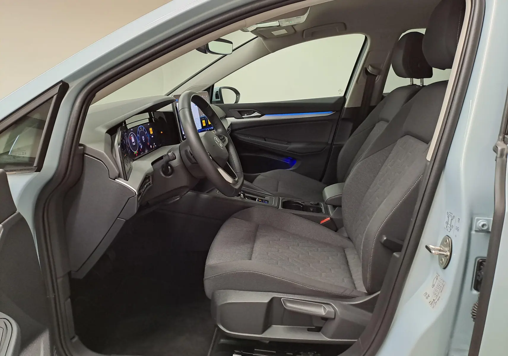Vue intérieure côté conducteur de la Volkswagen Golf 1.5 eTSI 2025 en Bleu Glacier, avec sièges tissu gris et éclairage d'ambiance bleu.