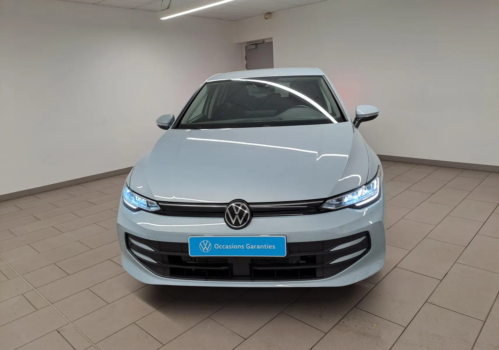 Vue avant d'une Volkswagen Golf 1.5 eTSI 116 DSG7 VW Edition en bleu glacier avec phares allumés dans un showroom.
