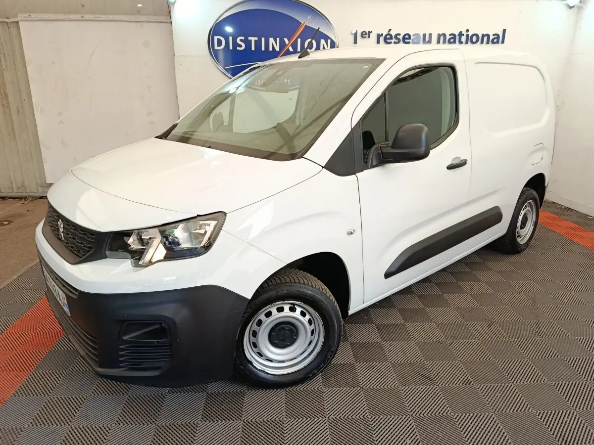 Vue 3/4 avant droit d’un Peugeot Partner blanc 2022 avec pare-chocs noirs et jantes acier en intérieur showroom.