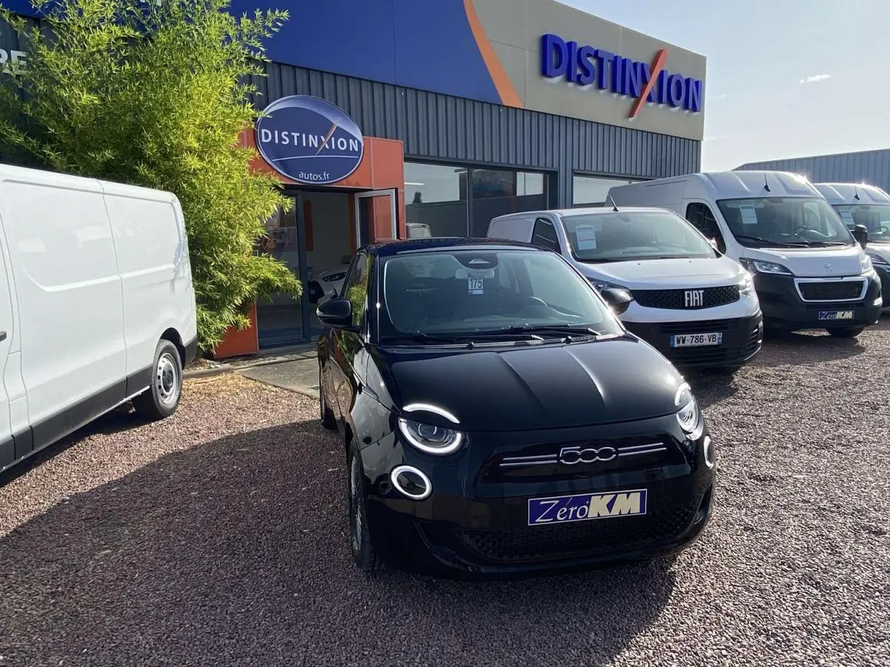 Vue avant d'une Fiat 500 électrique noire métal stationnée devant un concessionnaire avec d'autres véhicules blancs.