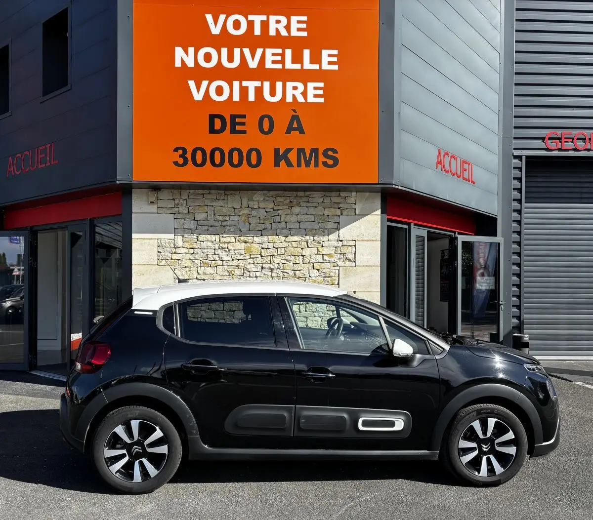 Profil droit d'une Citroën C3 noire avec toit blanc, garée devant un bâtiment avec panneau orange "Votre nouvelle voiture".