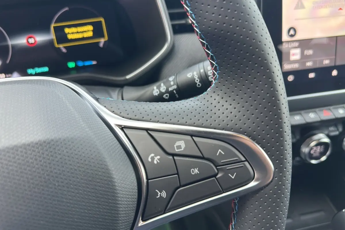 Gros plan sur le volant cuir perforé à commandes multifonctions de la Renault Clio Gris Rafale 2025, tableau de bord numérique en arrière-plan.