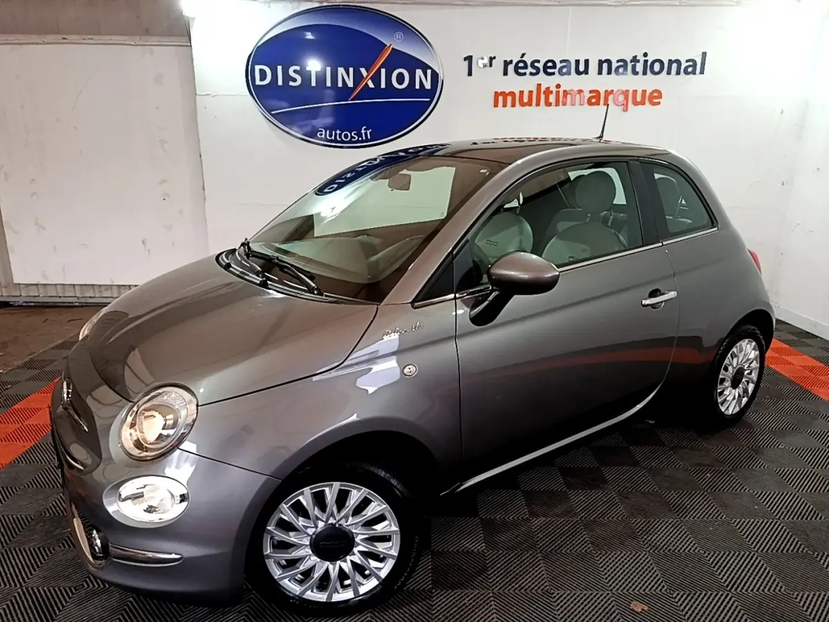 Fiat 500 Hybrid gris foncé vue 3/4 avant droit dans un showroom avec jantes argentées et phares allumés.