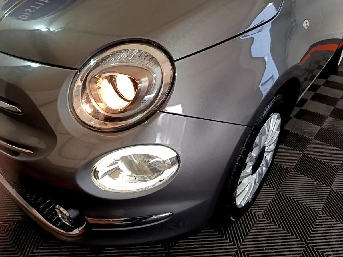 Gros plan sur l'avant droit gris foncé d'une Fiat 500 Hybrid avec phares allumés et jante alu visible.
