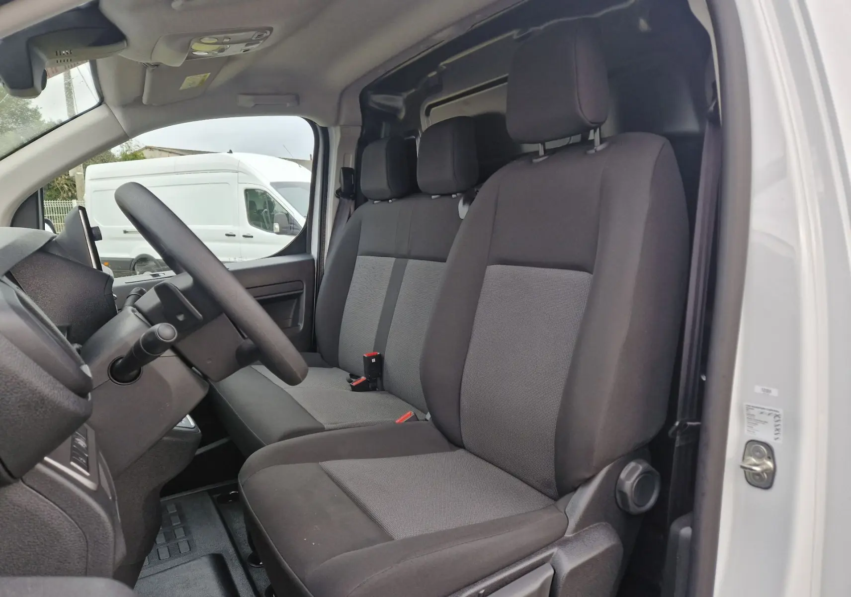 Vue intérieure côté conducteur du FIAT Scudo Fourgon blanc 2024, montrant les sièges tissu gris et noir, avec tableau de bord partiel.