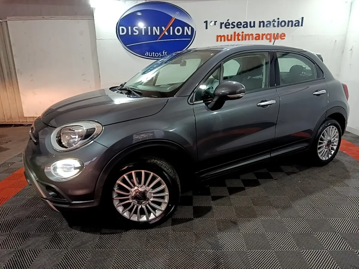 Fiat 500X gris foncé vue 3/4 avant droit, avec phares allumés et jantes alliage multibranches.